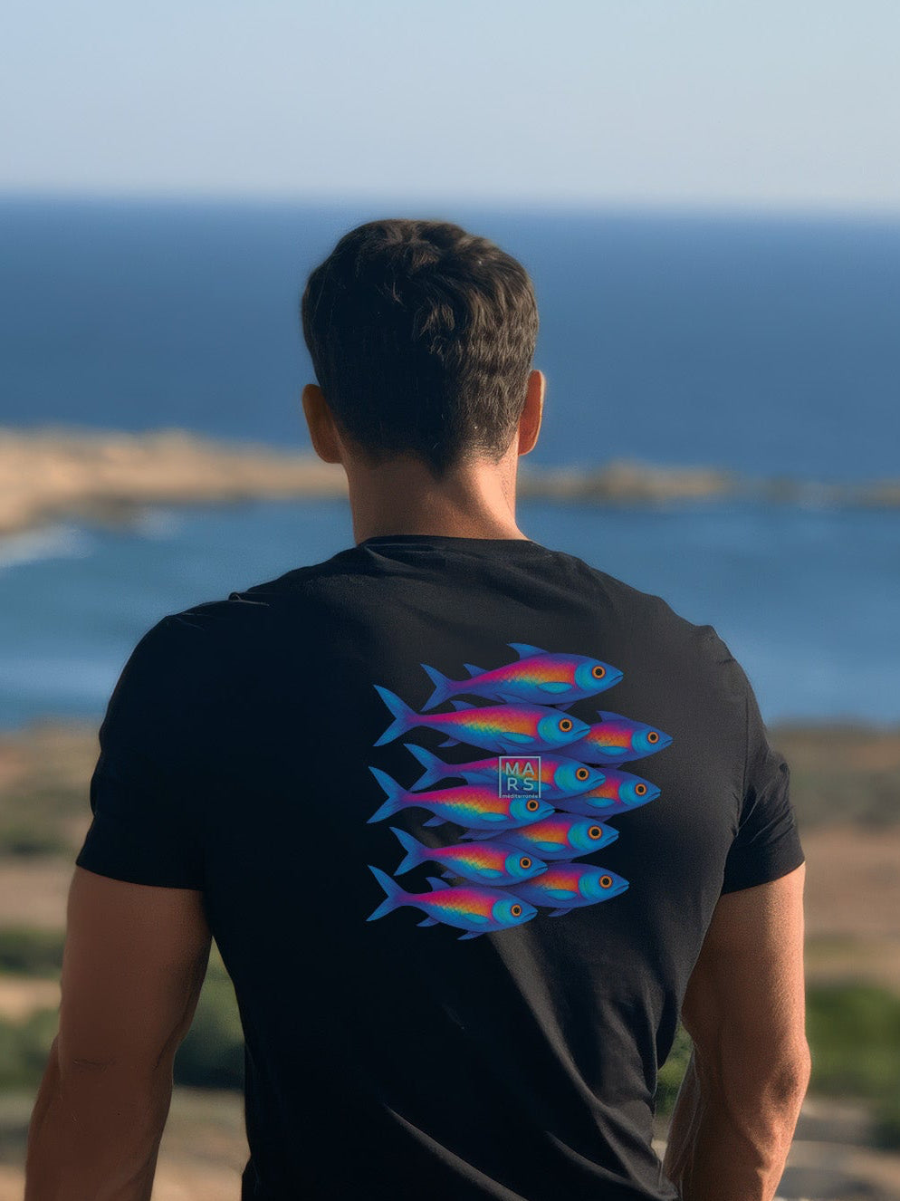 Homme au bord de mer portant un tee-shirt noir avec une illustration de sardines