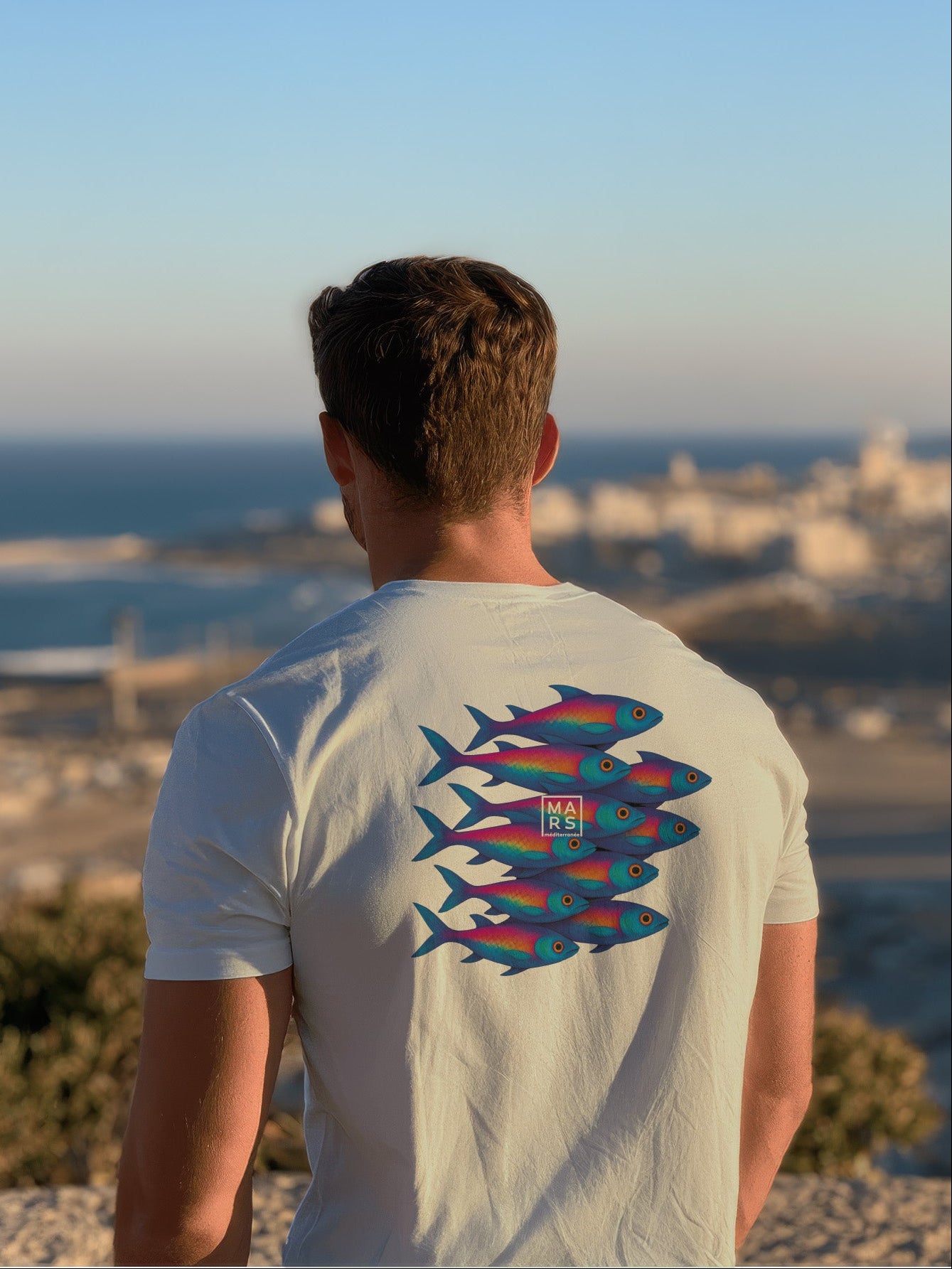 Homme au bord de mer portant un tee-shirt vert avec une illustration de sardines