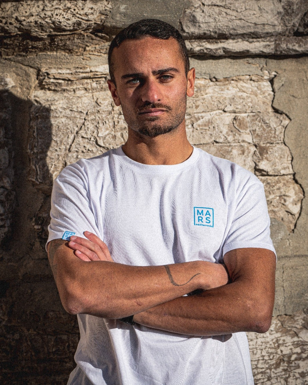 Un homme, les bras croisé, portant un T-Shirt blanc, chic et élégant, avec un logo bleu sur la poitrine