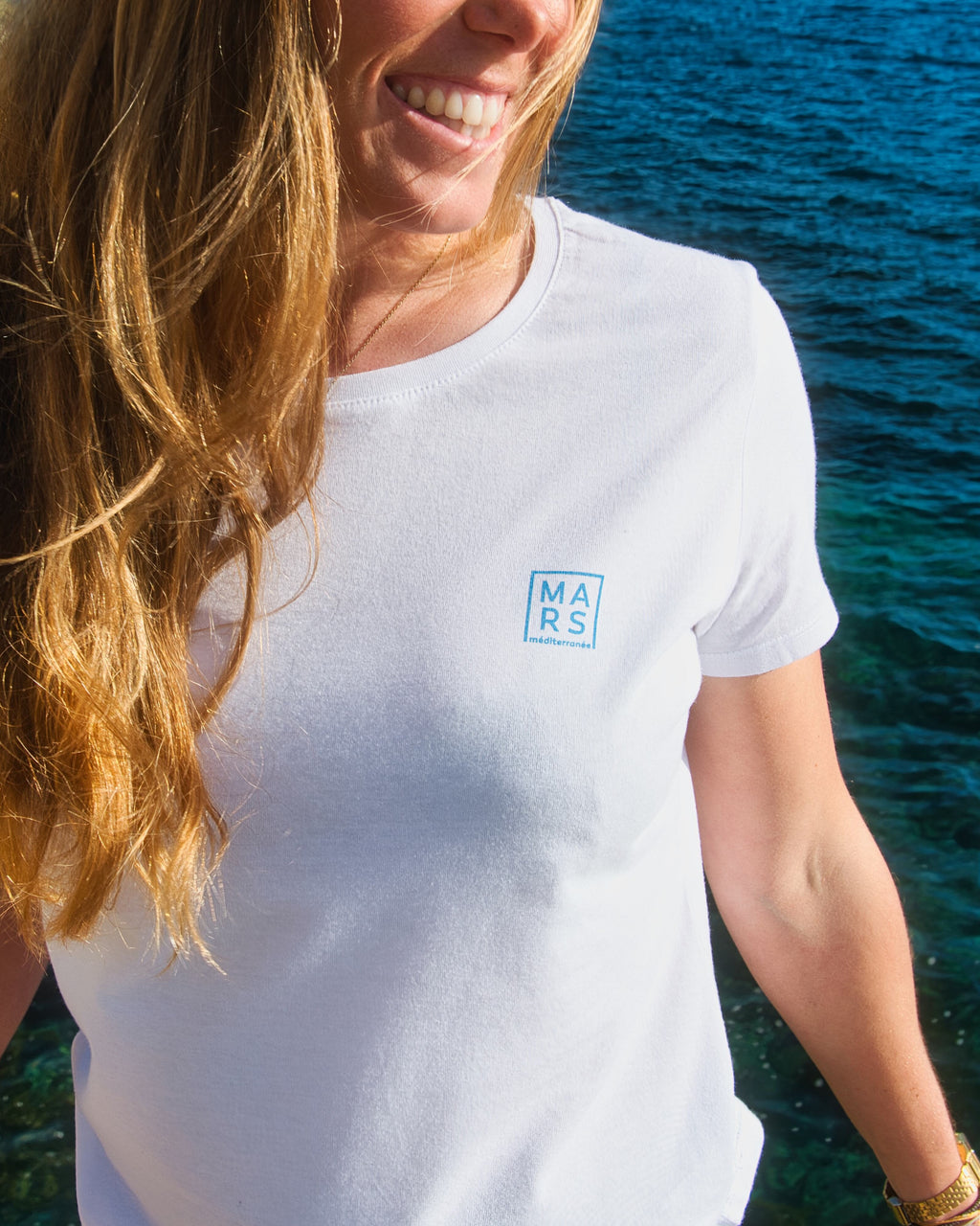 Femme au bord de mer portant un t-shirt blanc, très tendance, avec un logo MARS méditeranée en bleu