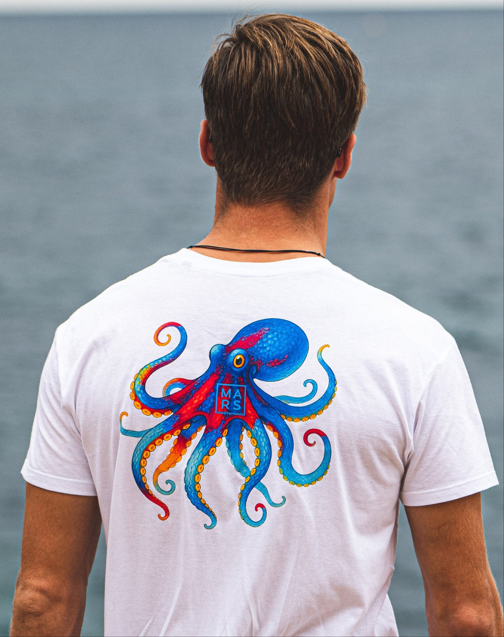 Un homme au bord de mer en portant un t-shirt blanc avec un motif de poulpe sur le dos
