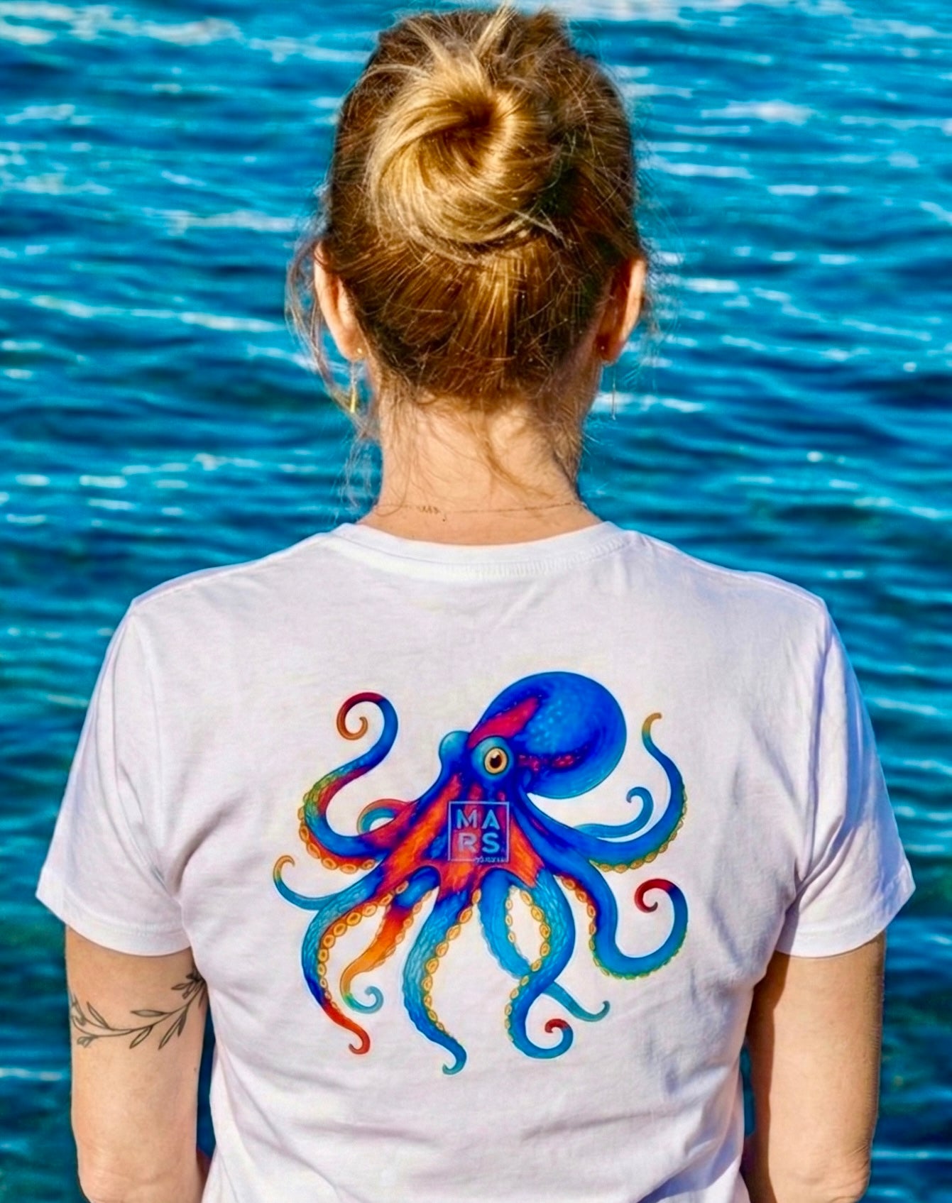 Une femme au bord de mer avec un T-Shirt blanc et une illustration de poulpe sur le dos