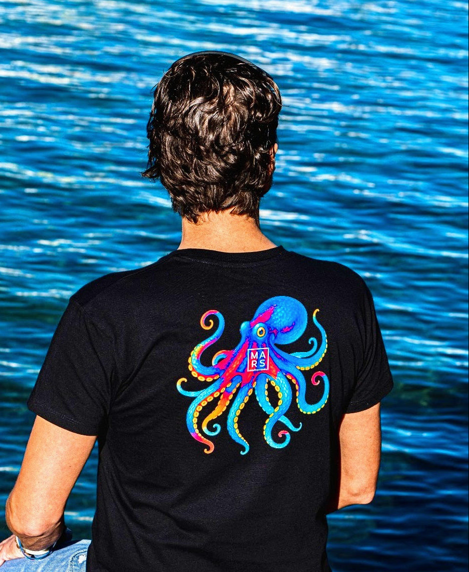 Homme en T-Shirt noir avec un illustration de poulpe sur le dos, au bord de la mer