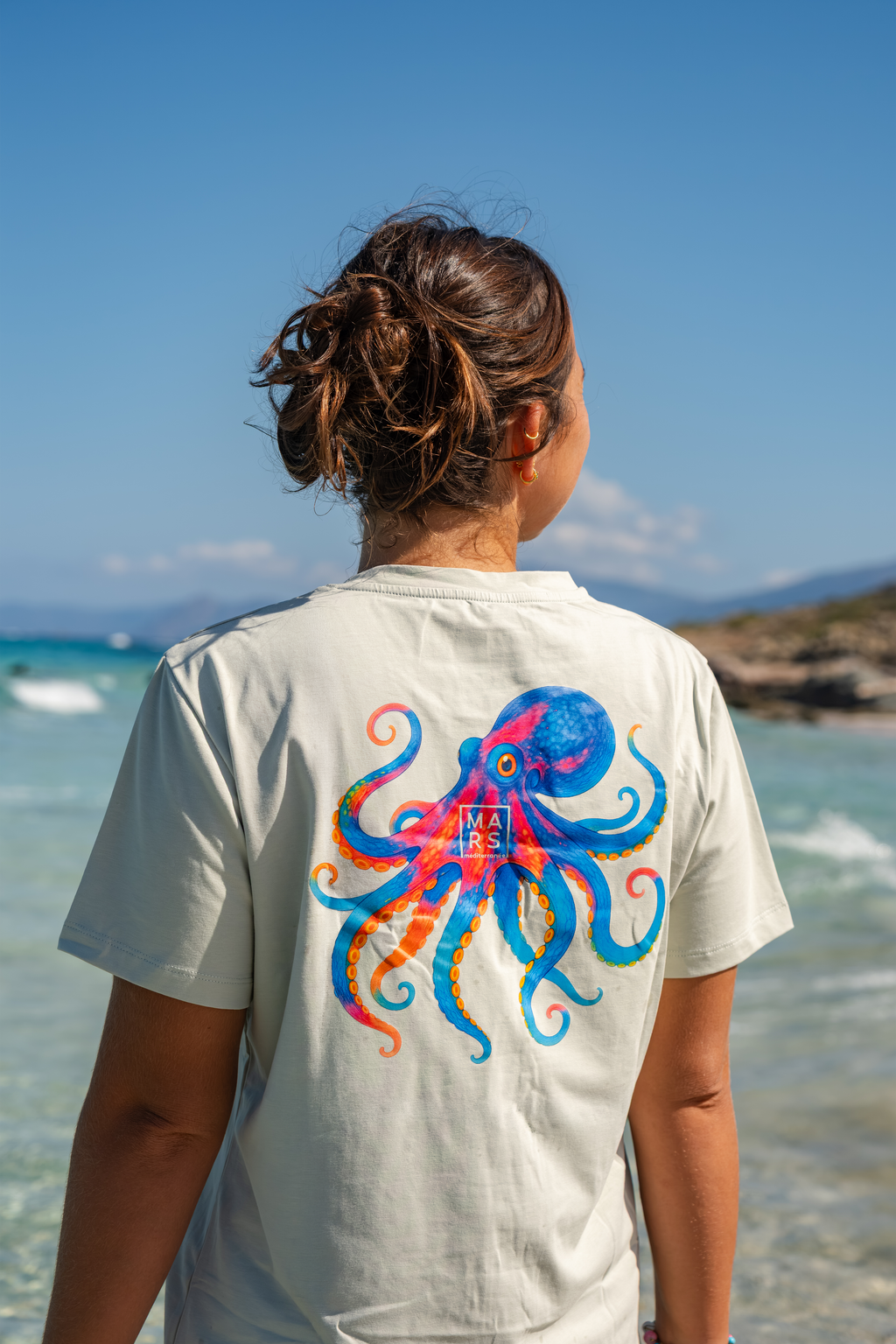 Une femme de dos, à la plage, portant un tee-shirt en vert sauge avec un joli motif de poulpe